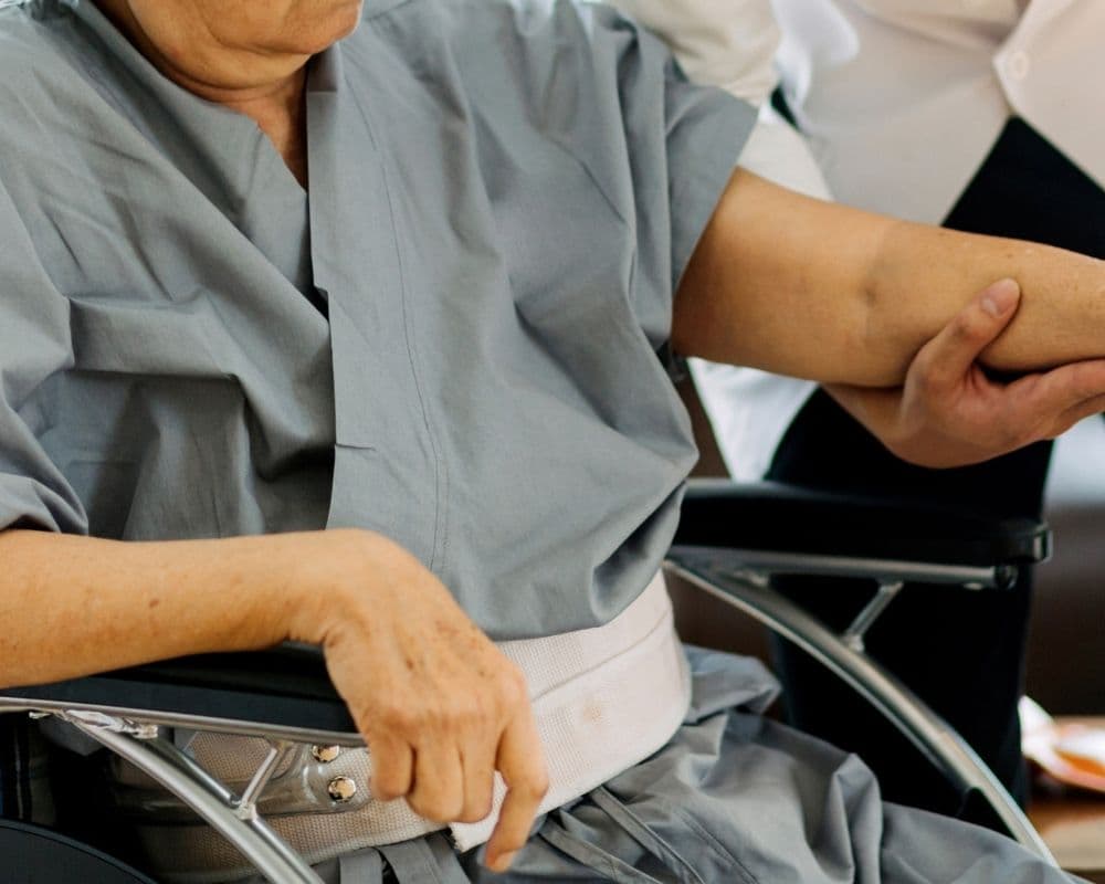 An elderly patient in a hospital gown seated in a wheelchair while a clinician gently supports their arm.